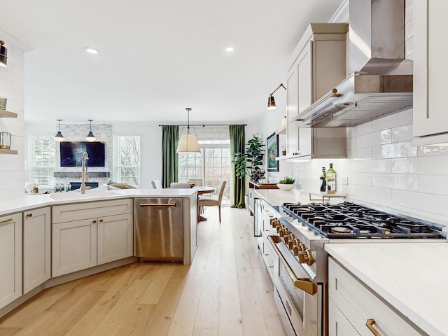 kitchen with appliances with stainless steel finishes, decorative light fixtures, backsplash, wall chimney range hood, and light hardwood / wood-style flooring