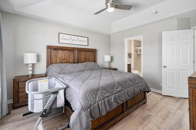 bedroom with a tray ceiling, ensuite bathroom, ceiling fan, and light wood-type flooring