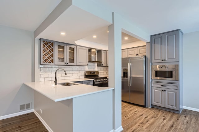 kitchen with wall chimney range hood, kitchen peninsula, sink, gray cabinetry, and stainless steel appliances