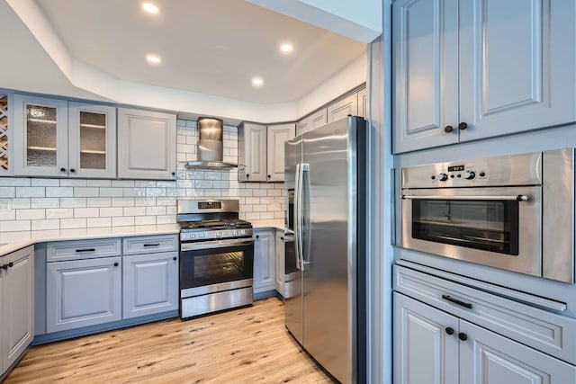 kitchen featuring stainless steel appliances, decorative backsplash, gray cabinetry, light wood-type flooring, and wall chimney exhaust hood