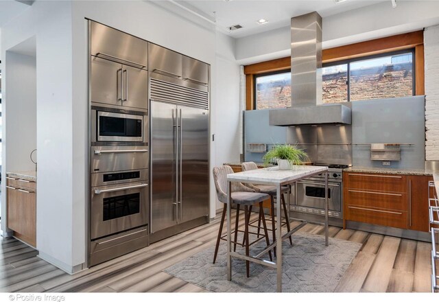 kitchen with built in appliances, tasteful backsplash, light hardwood / wood-style floors, island range hood, and light stone counters