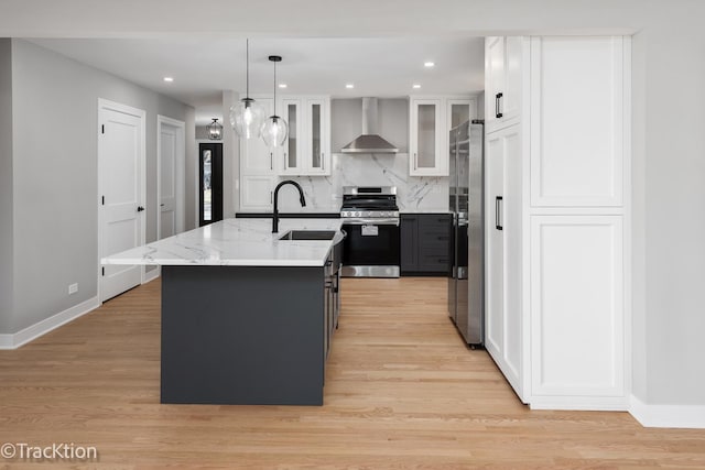 kitchen with white cabinetry, light wood-type flooring, appliances with stainless steel finishes, an island with sink, and wall chimney exhaust hood