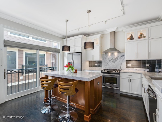 kitchen featuring wall chimney exhaust hood, appliances with stainless steel finishes, a kitchen island, pendant lighting, and white cabinets