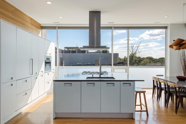 kitchen featuring light wood-type flooring, a kitchen breakfast bar, an island with sink, island exhaust hood, and oven