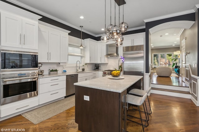 kitchen with built in appliances, white cabinets, a kitchen island, sink, and wall chimney exhaust hood