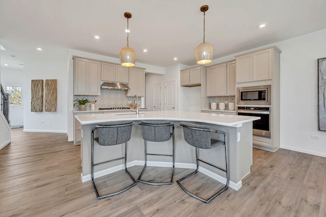 kitchen with pendant lighting, appliances with stainless steel finishes, an island with sink, backsplash, and light wood-type flooring