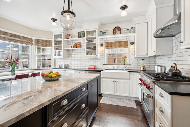 kitchen with wall chimney range hood, stainless steel stove, white cabinetry, dark stone countertops, and decorative light fixtures