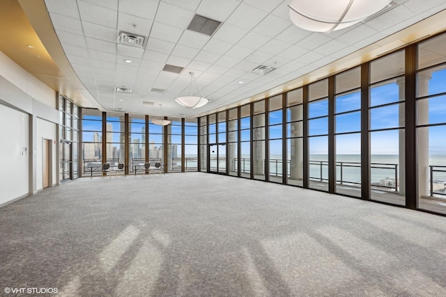 carpeted empty room featuring a towering ceiling, a paneled ceiling, a wall of windows, a water view, and a healthy amount of sunlight