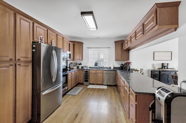 kitchen with light wood finished floors, appliances with stainless steel finishes, brown cabinetry, dark stone countertops, and beverage cooler