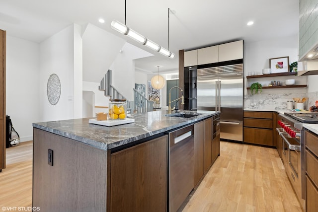 kitchen featuring hanging light fixtures, light wood-type flooring, dark stone counters, a center island with sink, and stainless steel appliances