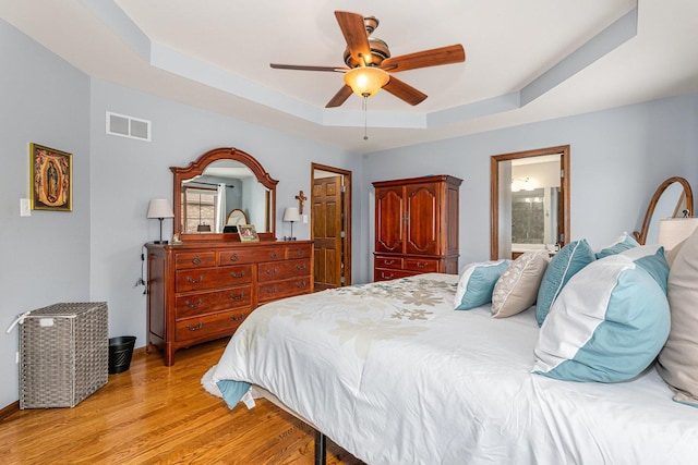 bedroom with light wood-type flooring, connected bathroom, ceiling fan, and a raised ceiling