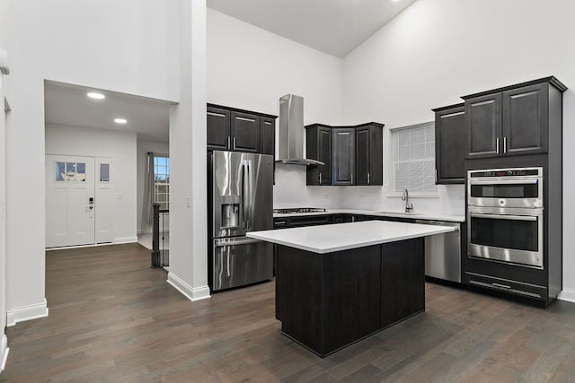 kitchen with appliances with stainless steel finishes, wall chimney exhaust hood, a towering ceiling, dark hardwood / wood-style floors, and a kitchen island