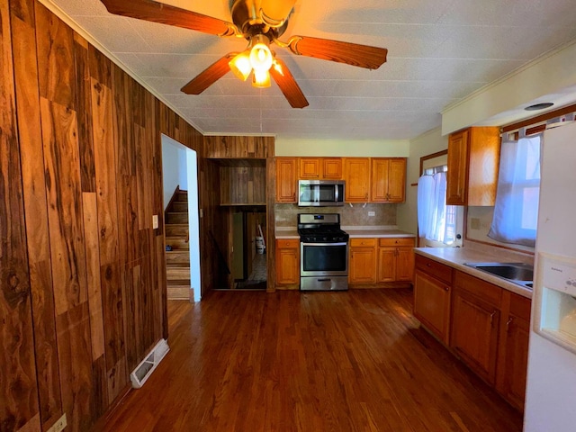 kitchen with ceiling fan, dark hardwood / wood-style flooring, sink, wood walls, and stainless steel appliances