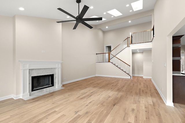 unfurnished living room featuring a towering ceiling, a skylight, ceiling fan, a premium fireplace, and light wood-type flooring