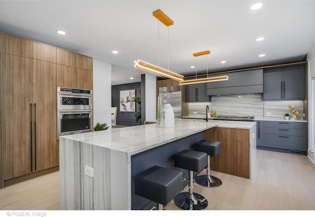 kitchen with appliances with stainless steel finishes, decorative light fixtures, a large island, light wood-type flooring, and light stone counters