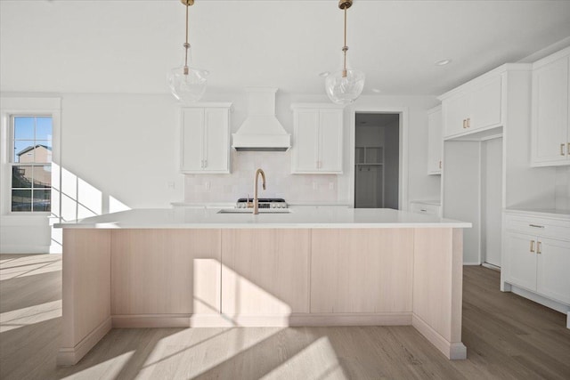 kitchen with pendant lighting, white cabinetry, custom range hood, and an island with sink