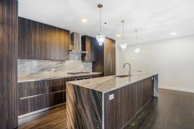 kitchen featuring wall chimney exhaust hood, sink, light stone counters, a center island with sink, and decorative backsplash