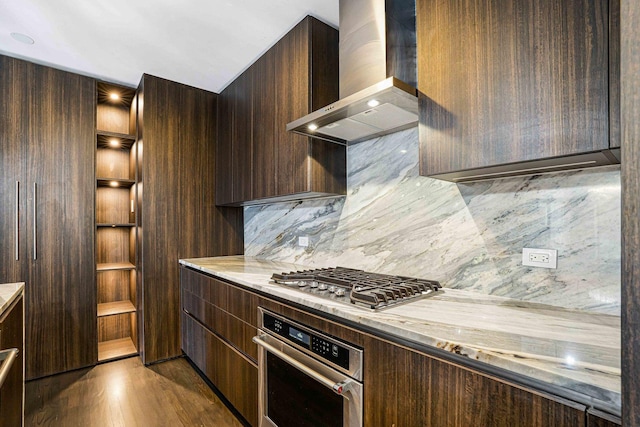 kitchen featuring dark brown cabinetry, wall chimney range hood, stainless steel appliances, tasteful backsplash, and light hardwood / wood-style flooring