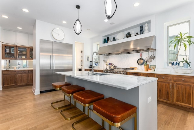 kitchen with sink, built in refrigerator, exhaust hood, light hardwood / wood-style floors, and a center island with sink