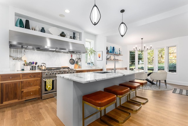 kitchen with light stone counters, double oven range, pendant lighting, a kitchen island with sink, and exhaust hood