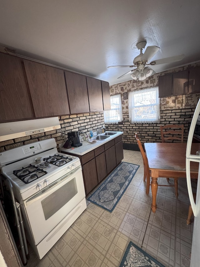 kitchen featuring sink, gas range gas stove, ceiling fan, dark brown cabinetry, and brick wall