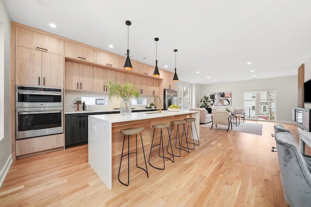 kitchen with stainless steel appliances, hanging light fixtures, a center island with sink, and light brown cabinets