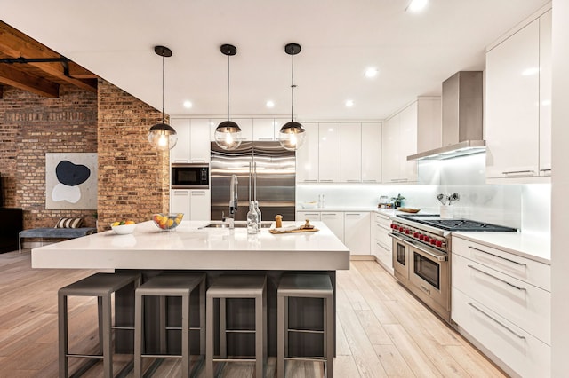 kitchen with wall chimney exhaust hood, built in appliances, an island with sink, brick wall, and white cabinets