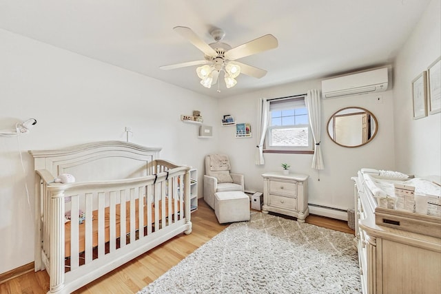 bedroom with a wall mounted AC, light wood-type flooring, a baseboard radiator, a nursery area, and ceiling fan