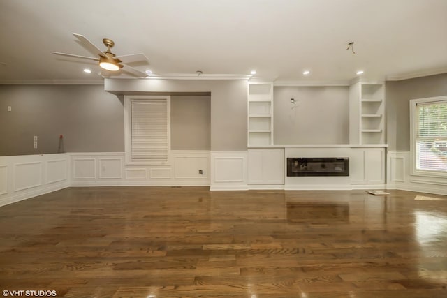 unfurnished living room featuring built in shelves, dark wood-type flooring, ornamental molding, and ceiling fan