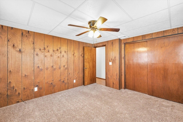 unfurnished bedroom featuring a drop ceiling, wood walls, ceiling fan, and carpet flooring