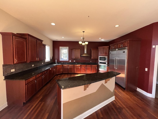 kitchen with wall chimney exhaust hood, built in appliances, hanging light fixtures, dark hardwood / wood-style flooring, and a kitchen island