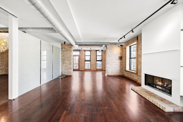 unfurnished living room featuring track lighting, wood finished floors, brick wall, and a lit fireplace