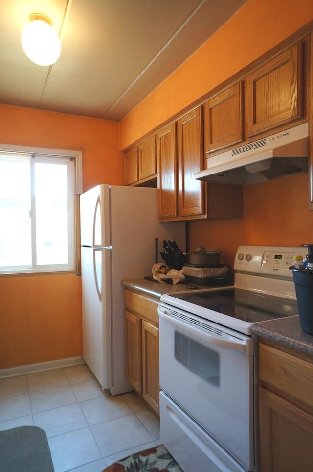 kitchen featuring white appliances, light tile patterned floors, brown cabinetry, under cabinet range hood, and dark countertops