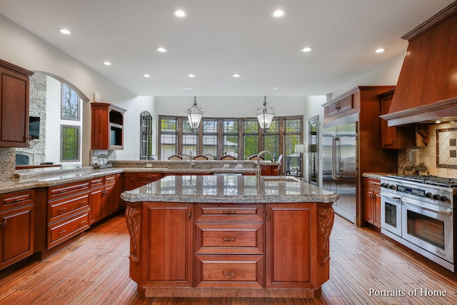 kitchen with sink, backsplash, high end appliances, a center island with sink, and light wood-type flooring