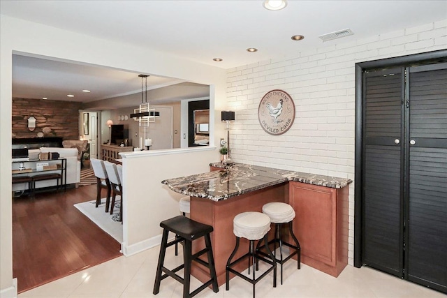 kitchen with dark stone countertops, a kitchen bar, brick wall, decorative light fixtures, and kitchen peninsula