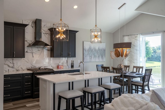 kitchen with hanging light fixtures, backsplash, stainless steel gas range oven, a center island with sink, and wall chimney exhaust hood