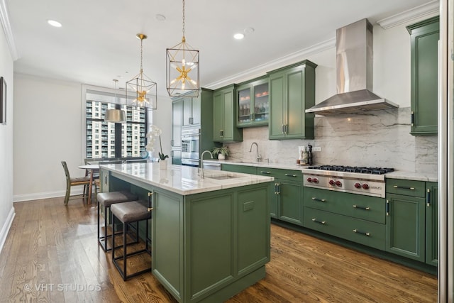 kitchen with green cabinetry, an island with sink, appliances with stainless steel finishes, and wall chimney exhaust hood