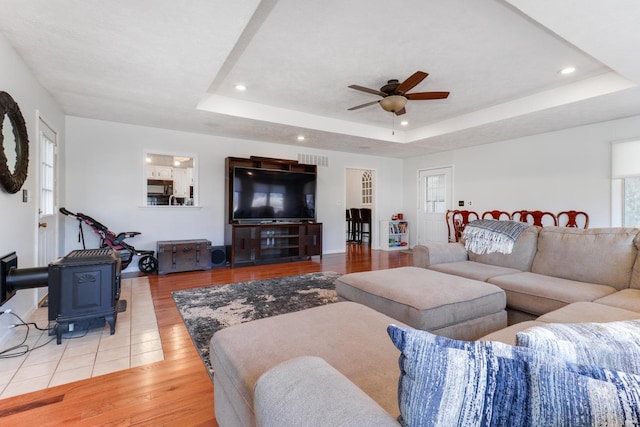 living room featuring light hardwood / wood-style flooring, a raised ceiling, ceiling fan, and a wood stove