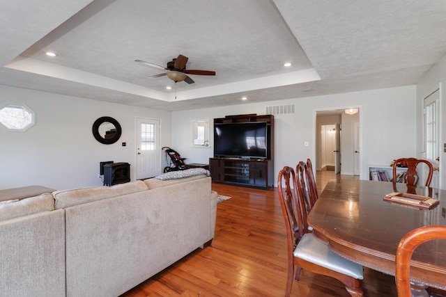 living room featuring a textured ceiling, a wood stove, light wood-type flooring, a raised ceiling, and ceiling fan