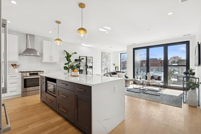 kitchen featuring decorative light fixtures, light hardwood / wood-style floors, stainless steel appliances, wall chimney exhaust hood, and white cabinets