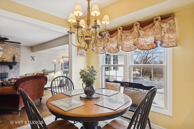 dining room featuring wood-type flooring, a brick fireplace, ceiling fan with notable chandelier, and beam ceiling