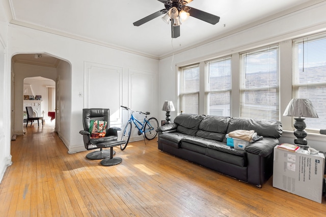 living area with arched walkways, light wood-style floors, crown molding, and a ceiling fan