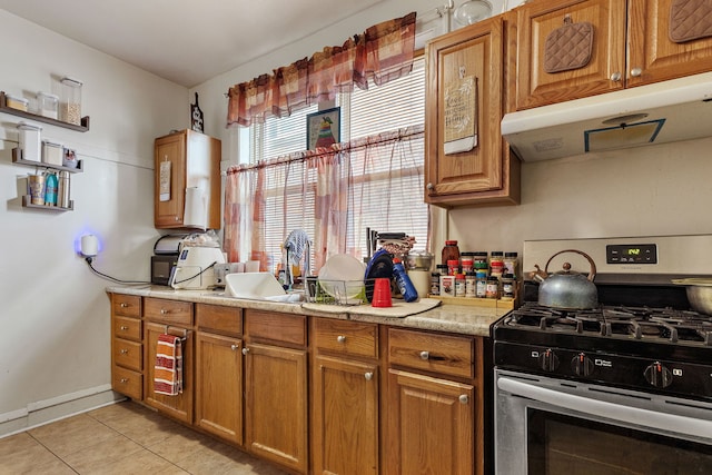 kitchen featuring light tile patterned floors, gas stove, light countertops, under cabinet range hood, and brown cabinets