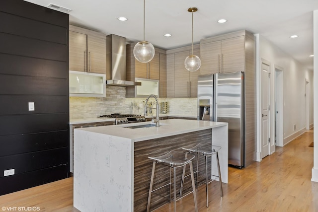 kitchen with pendant lighting, sink, stainless steel appliances, a center island with sink, and wall chimney range hood