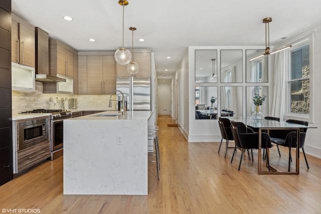 kitchen featuring sink, appliances with stainless steel finishes, pendant lighting, a kitchen island with sink, and wall chimney range hood