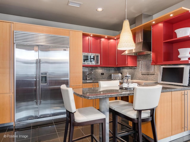 kitchen featuring wall chimney range hood, sink, backsplash, built in appliances, and dark tile patterned flooring