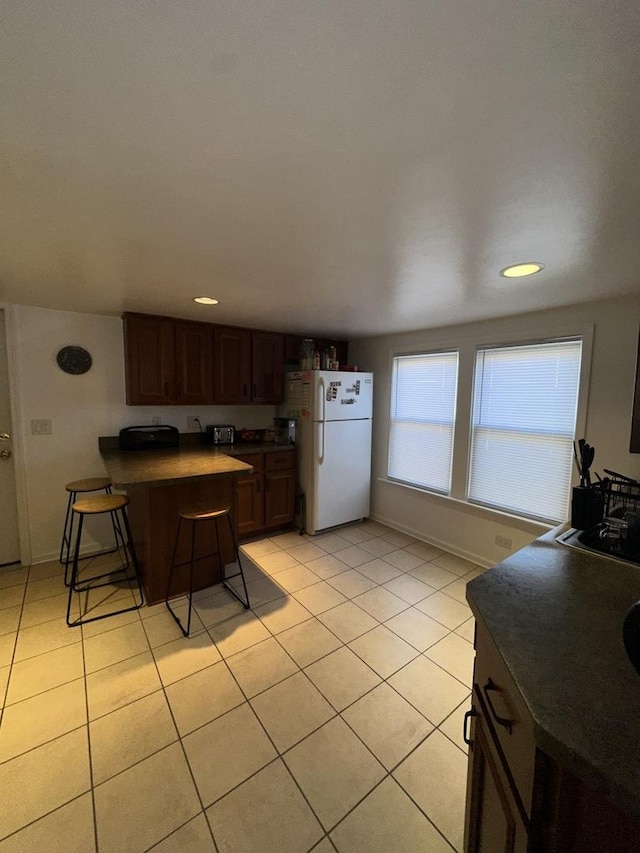 kitchen with light tile patterned floors, dark brown cabinets, a kitchen breakfast bar, white refrigerator, and kitchen peninsula