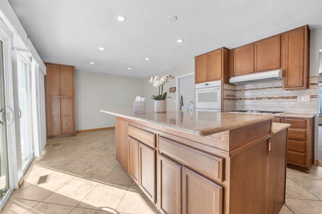 kitchen with brown cabinetry, a kitchen island with sink, under cabinet range hood, stainless steel gas cooktop, and backsplash