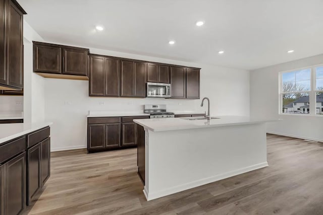 kitchen featuring appliances with stainless steel finishes, sink, a kitchen island with sink, dark brown cabinets, and light wood-type flooring