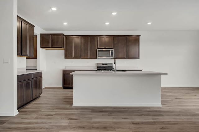 kitchen with sink, a kitchen island with sink, dark brown cabinetry, stainless steel appliances, and light wood-type flooring